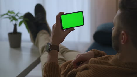 man-is-holding-smartphone-horizontal-and-viewing-video-resting-at-home-or-office-green-screen-technology-for-montage-modern-gadget-and-application